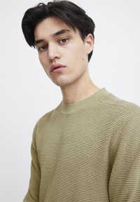 Pulover tricotat de culoare verde măsliniu, cu un model texturat, guler rotund și o bandă subțire pe guler și pe manșete.