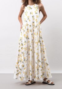 En model bærer en hvid, blomsteret maxikjole med gule og grønne mønstre, parret med lyse sandaler, mens hun står foran en neutral baggrund.