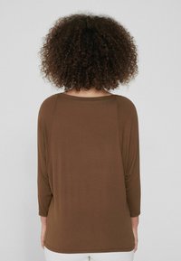 Braunes Langarmshirt mit lockerer Passform, rundem Halsausschnitt und Dreiviertelärmeln. Glatte Textur ohne sichtbare Muster.