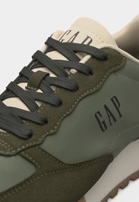 Αθλητικό παπούτσι χαμηλού κοψίματος με πάνελ από πράσινο σουέντ και ύφασμα, σκούρα γκρι κορδόνια και λογότυπο "GAP" στο πλάι και στη γλώσσα.