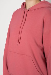 Mikina s kapucí v barvě zaprášené růže s nastavitelným šňůrkovým stahováním, přední kapsou a žebrovanými manžetami. Vyrobena z měkké látky s pohodlným střihem.