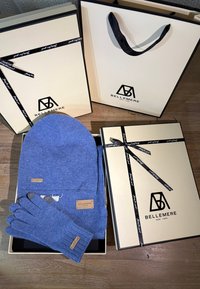 Bonnet et gants tricotés bleus présentés avec des boîtes crème ornées de rubans noirs et de logos. Les boîtes sont soigneusement disposées, mettant en valeur une présentation de cadeau.