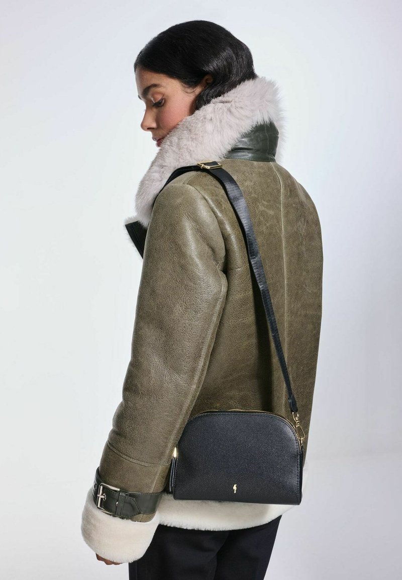 Oliwkowa skórzana kurtka z miękkim beżowym futerkiem na kołnierzu, zestawiona z czarną torebką na ramię z złotym zamkiem i akcentami.