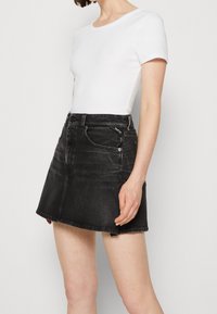 Černá denimová sukně s mírně do zvonu se rozšiřujícím tvarem, pětikapsovým designem a viditelným prošitím. Kombinována s přiléhavým bílým tričkem.