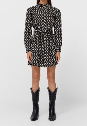 Femme portant une robe boutonnée à motif ondulé noir et blanc avec une ceinture à la taille et des bottes de cowboy noires, debout devant un fond uni.