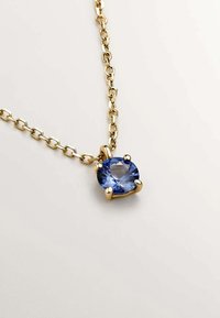 Collar de cadena de oro que presenta una gran gema azul redonda engastada en cuatro garras, mostrando una superficie lisa y variaciones de color claras.