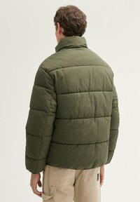 Grøn dunjakke med quiltet mønster, høj krave og ribbet detaljer ved manchetterne, båret over beige bukser. Glat tekstur, afslappet design.