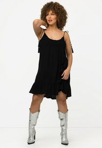 Vestido negro sin mangas con un dobladillo con volantes, que presenta tirantes ajustables y un corpiño texturizado. La modelo lleva botas de rodilla plateadas y brillantes.