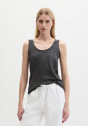 Jeune femme aux longs cheveux blonds portant un haut sans manches gris foncé et un pantalon blanc à cordon, debout devant un fond blanc uni.