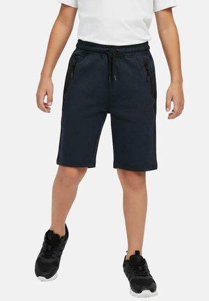 Kind draagt zwarte sportbroek met ritszakken, wit T-shirt en zwarte sneakers, staat met licht gespreide benen.