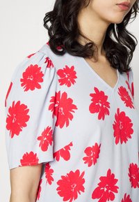 Blusa azul clara com padrões florais vermelhos ousados, mangas curtas e bufantes, e decote em V, feita de um tecido suave e texturizado.