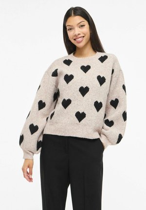 Hellrosa Pullover mit schwarzen Herzmustern, langen Puffärmeln und einem gerippten Ausschnitt. Trägt dazu eine schwarze Hose.
