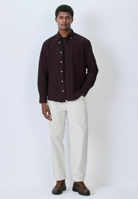 Dunkelburgunderfarbendes Hemd mit Knöpfen, schwarzes T-Shirt darunter, hellbeige Hose, braune Schnürstiefel mit orangen Akzenten, vor einem einfarbigen Hintergrund stehend.