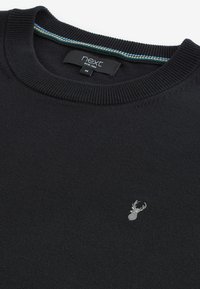 Pulover negru din tricot cu decolteu rotund, având un mic logo brodat cu un cerb argintiu pe piept și o dublură colorată în interiorul gulerului.