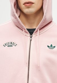 Personne portant un sweat à capuche zippé Adidas Originals rose clair avec la capuche relevée, un t-shirt noir visible en dessous, et une broderie de logo verte.