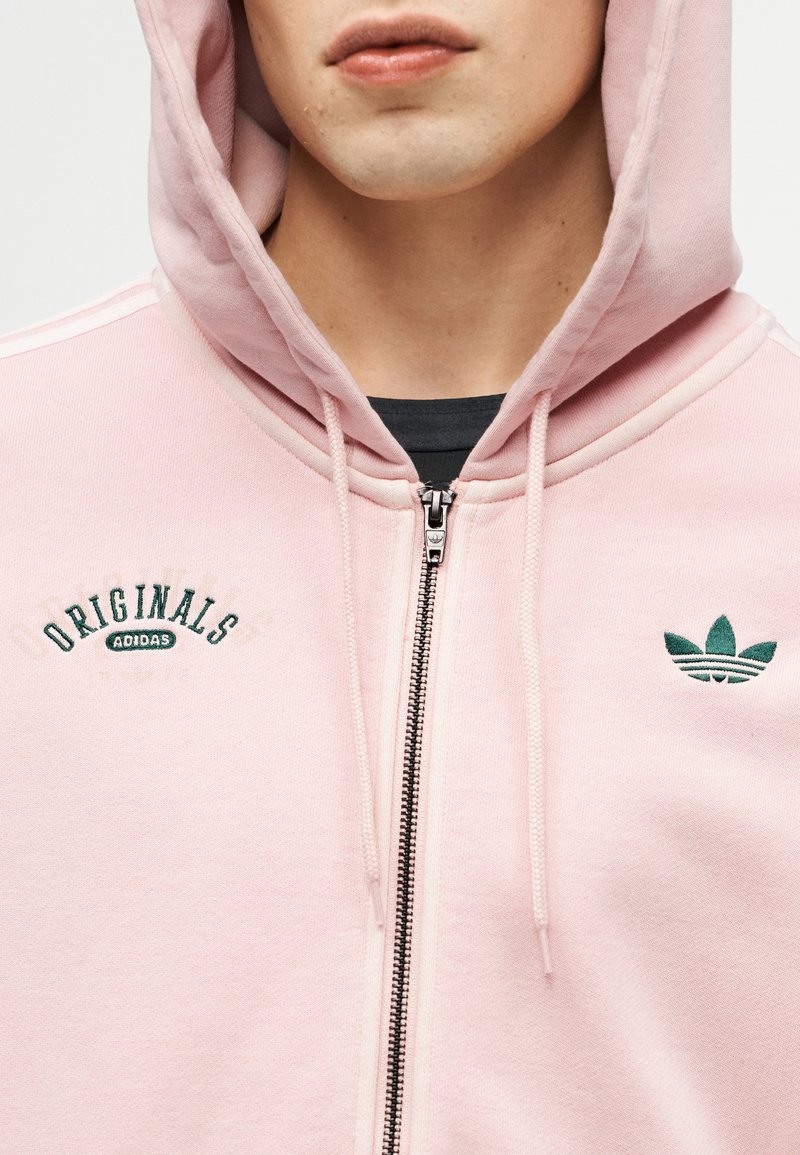 Personne portant un sweat à capuche zippé Adidas Originals rose clair avec la capuche relevée, un t-shirt noir visible en dessous, et une broderie de logo verte.