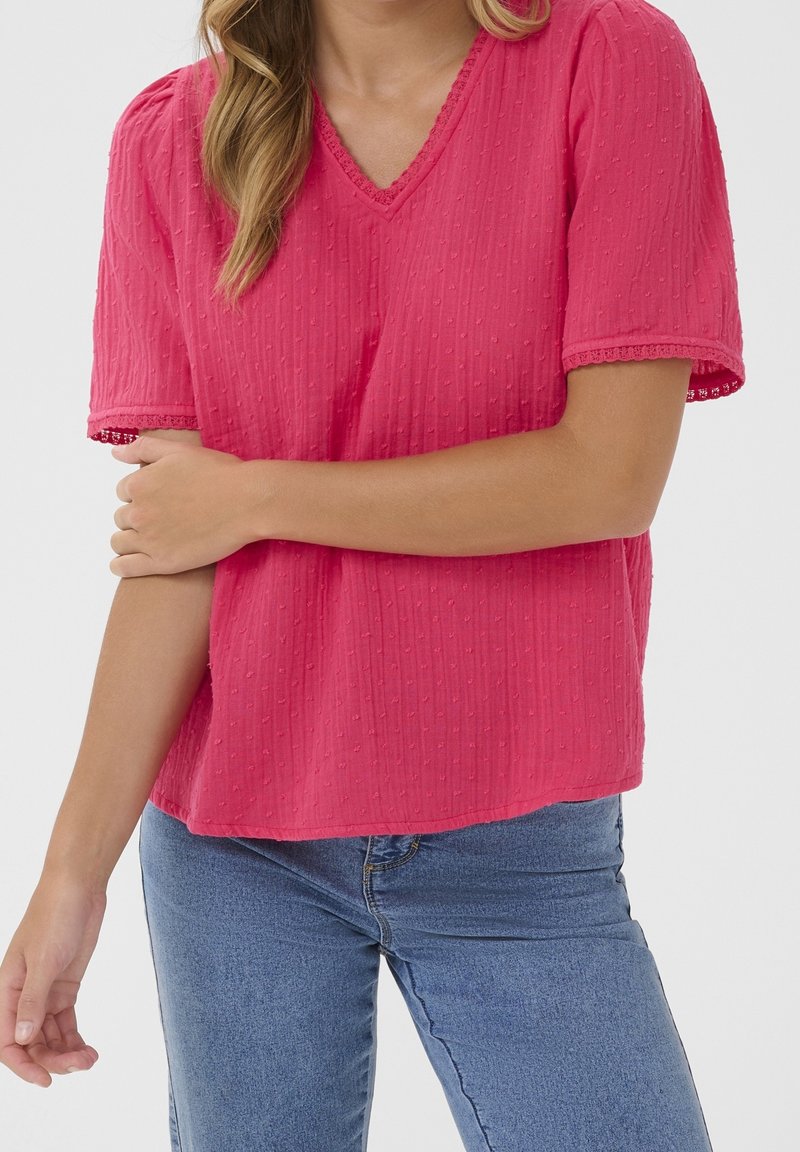 Femme portant un haut rose vif texturé à manches courtes avec bordure en dentelle et un jean bleu, tenant un bras croisé sur son corps.