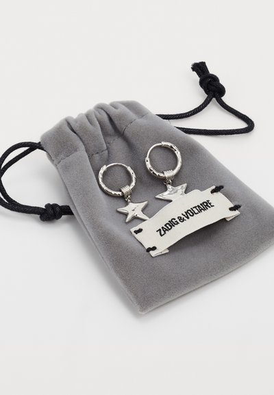Hopeiset rengaskorvakorut tähtikiinnikkeillä, jotka lepäävät harmaassa samettipussissa. Pussissa on valkoinen etiketti mustalla tekstillä, jossa lukee "Zadig & Voltaire."