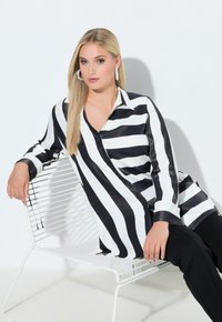 Černobílá proužkovaná halenka s límečkem, dlouhými rukávy a volným střihem, kombinovaná s černými kalhotami, sedící na bílé drátěné židli.