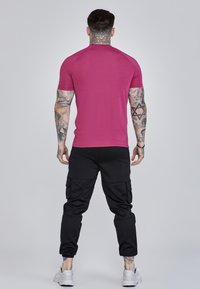 Īsa piedurkņu, pieguļošs t-kreklis spilgtā rozā krāsā, kombinēts ar melnām armijas biksēm ar sānu kabatām. Modelis stāv ar muguru pret skatītāju, demonstrējot apģērba formas.