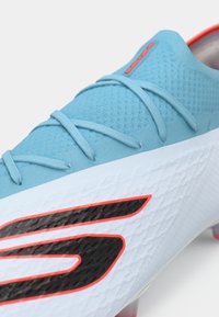 Chuteira de futebol turquesa com um material texturizado, apresentando acabamentos em preto e vermelho, um design contornado e um sistema de atilhos visível.