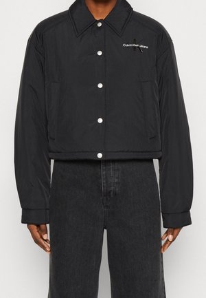 Black cropped button-up jacket with Calvin Klein Jeans logo on left chest, worn with high-waist dark gray jeans.