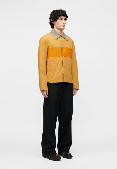 Άνδρας όρθιος, φορώντας ένα μουσταρδί κίτρινο σακάκι με κουμπιά, με πορτοκαλί ρίγα και ελιά για γιακά, μαύρο παντελόνι φαρδύ και σκούρα παπούτσια.