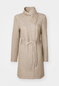 Beige pitkä takki, joka on tehty teksturoidusta kankaasta. Siinä on korkea kaulus, epäsymmetrinen etusuljin ja vyö, joka sopii vyötärölle.