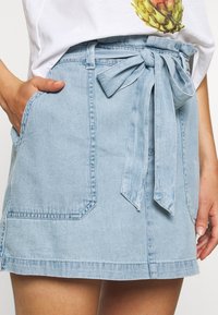 Světle modrá džínová sukně s páskem na zavázání v pase, dvěma předními kapsami a mírně rozšířeným lemem. Materiál působí texturovaně a neformálně.