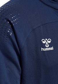 Mörkblå atletisk tyg med texturerad yta, med en gradient av glänsande prickar och en vit logotyp av "hummel" som står ut.