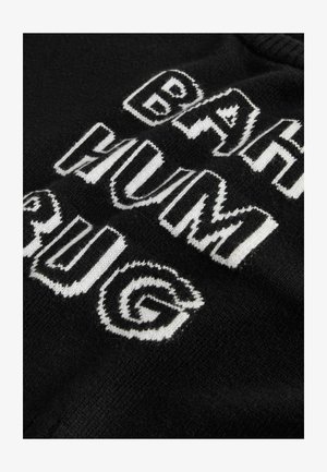 Melnā adītā džemperī ir izteiksmīgs balts uzraksts "BAH HUM BUG" ar lielajiem burtiem un stilizētu rakstu. Ir redzama teksturēta auduma detaļa.