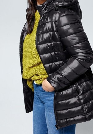 Femme portant une doudoune matelassée noire par-dessus un gros pull vert et un jean bleu, avec une main dans la poche.