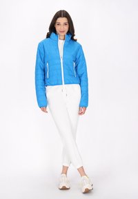 Jachetă albastră izolată cu guler în picioare, fermoar alb și două buzunare laterale, purtată peste pantaloni albi tapered și sneakers ușori.