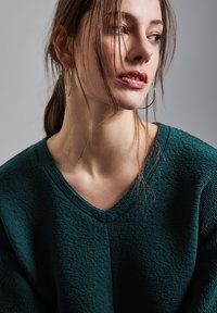 Maglione verde con texture e scollatura a V, indossato da un modello. Capelli acconciati con ciocche sciolte, che completano grandi orecchini a cerchio in argento.