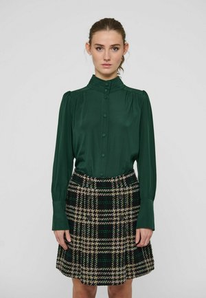 Kvinde iført en mørkegrøn bluse med knapper og høj krave samt et sort, beige og grønt ternet skørt med dekorative knapper.