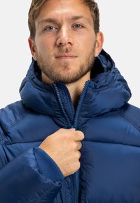 Mladý muž s svetlou bradou zapína modrú páperovú bundu s kapucňou, čelom k prednej strane na jednoduchom pozadí.