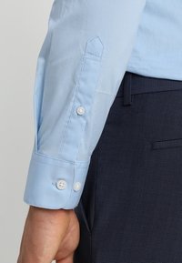 Camicia azzurra chiara con polsini a punta, dotata di due bottoni bianchi, contrastata da pantaloni scuri con texture. Il tessuto è liscio.