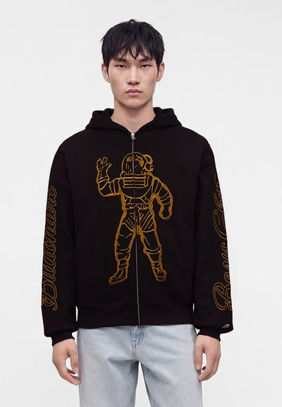 Young man wearing a black hoodie with a large gold astronaut design on front and gold text on sleeves, paired with light blue jeans.