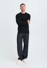 Zwarte gebreide trui, relaxed-fit donkerdenim jeans en witte sneakers. Model staat met gekruiste armen tegen een lichte achtergrond.