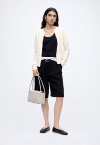 Femme portant un cardigan blanc sur un haut noir, un short noir longueur genou, des ballerines noires, tenant un petit sac beige.