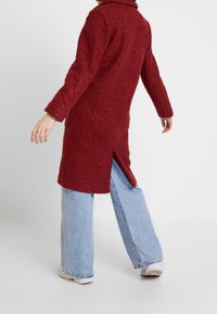 Abrigo largo rojo texturizado con cuello, aberturas laterales y ajuste holgado, combinado con vaqueros de pierna ancha en azul claro y zapatillas blancas.