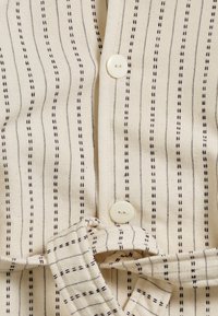 Vaalean beige kangas, jossa on ohuita tummia raidoituksia, varustettuna napilla suljettavalla ja sidotulla kangasdetaililla. Rakenne on teksturoitu, kudottu materiaali.