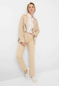 Beige cotton blazer with a notched collar, white button-up shirt, beige fitted pants, and white sneakers. Model poses with one hand on hip.