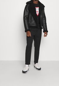 Schwarze Lederjacke mit einem Fellkragen, getragen über einem schwarzen T-Shirt mit einem weißen und roten Grafikmotiv, kombiniert mit schwarzen Hosen und Sneakers.