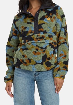 Kvinde iført flerfarvet camouflage fleecepullover med sorte trykknapper og blå jeans, hænder afslappet ved siderne.