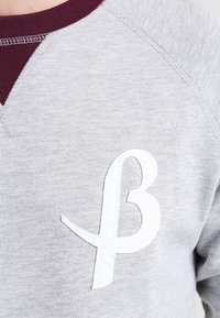 Felpa grigia con colletto maroon, caratterizzata da un logo a lettera "B" bianco e testurizzato sul petto. Tessuto morbido con un design informale.