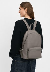 Mochila de cuero gris con una textura suave, forma redondeada, bolsillo frontal con cremallera y correas ajustables, llevada en la espalda.