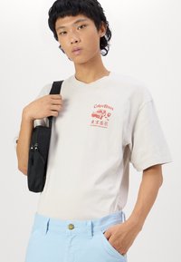 Camiseta de algodón beige con gráfico de "Tokyo Roses" en rojo. Combinada con pantalones azul claro y un bolso cruzado negro. Diseño sencillo y ajuste casual.