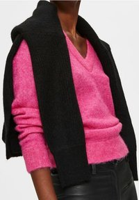 Iemand draagt een levendig roze trui met een diepe V-hals en een zwarte cardigan die over hun schouders is gedrapeerd, in combinatie met donkere broek.