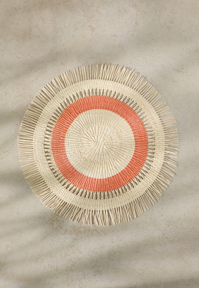 Round woven placemat with a fringed edge. Features concentric circles in cream and coral. Made of natural fibers, textured surface.
